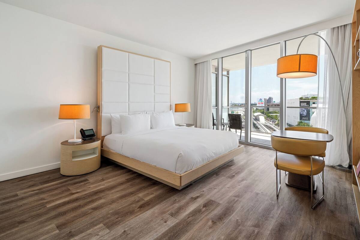 Een moderne hotelkamer met een groot bed, twee nachtkastjes met lampen, een kleine tafel en stoel, en een groot raam met uitzicht op de stad.