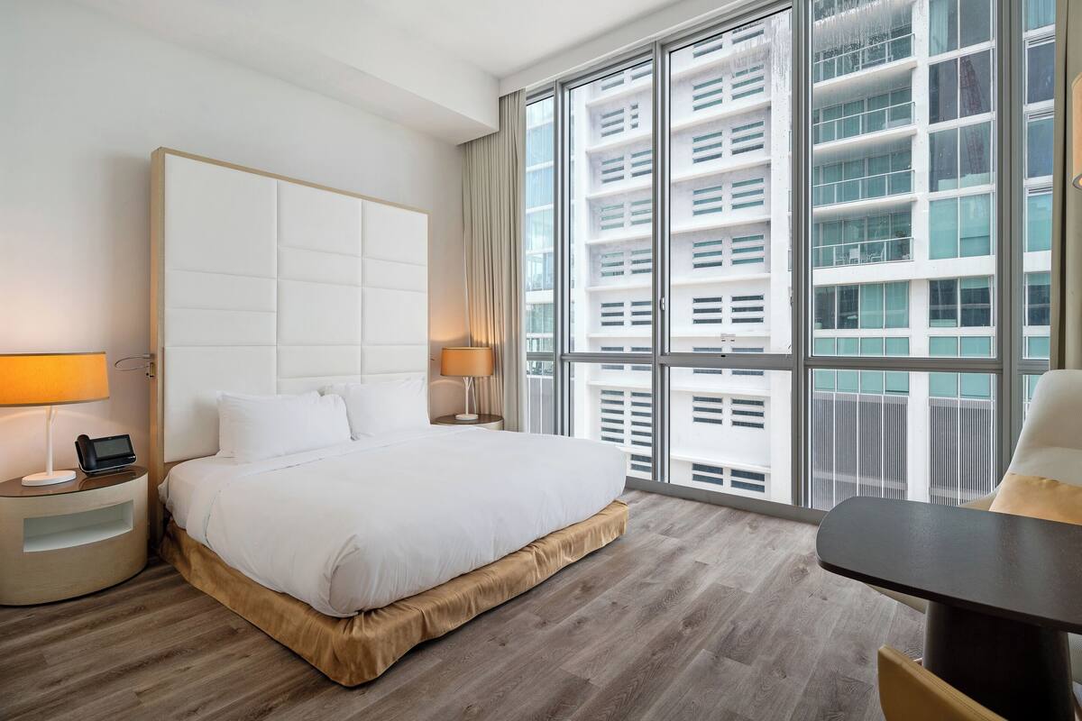 Een moderne hotelkamer met een groot bed, nachtkastjes, een bureau en een groot raam met uitzicht op de stad.