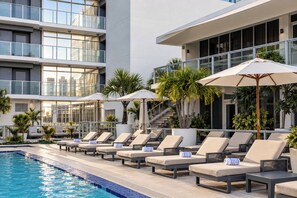 A poolside area with lounge chairs and umbrellas, a clear pool, and a modern building with large windows. - The Grayson Miami Downtown (Miami)