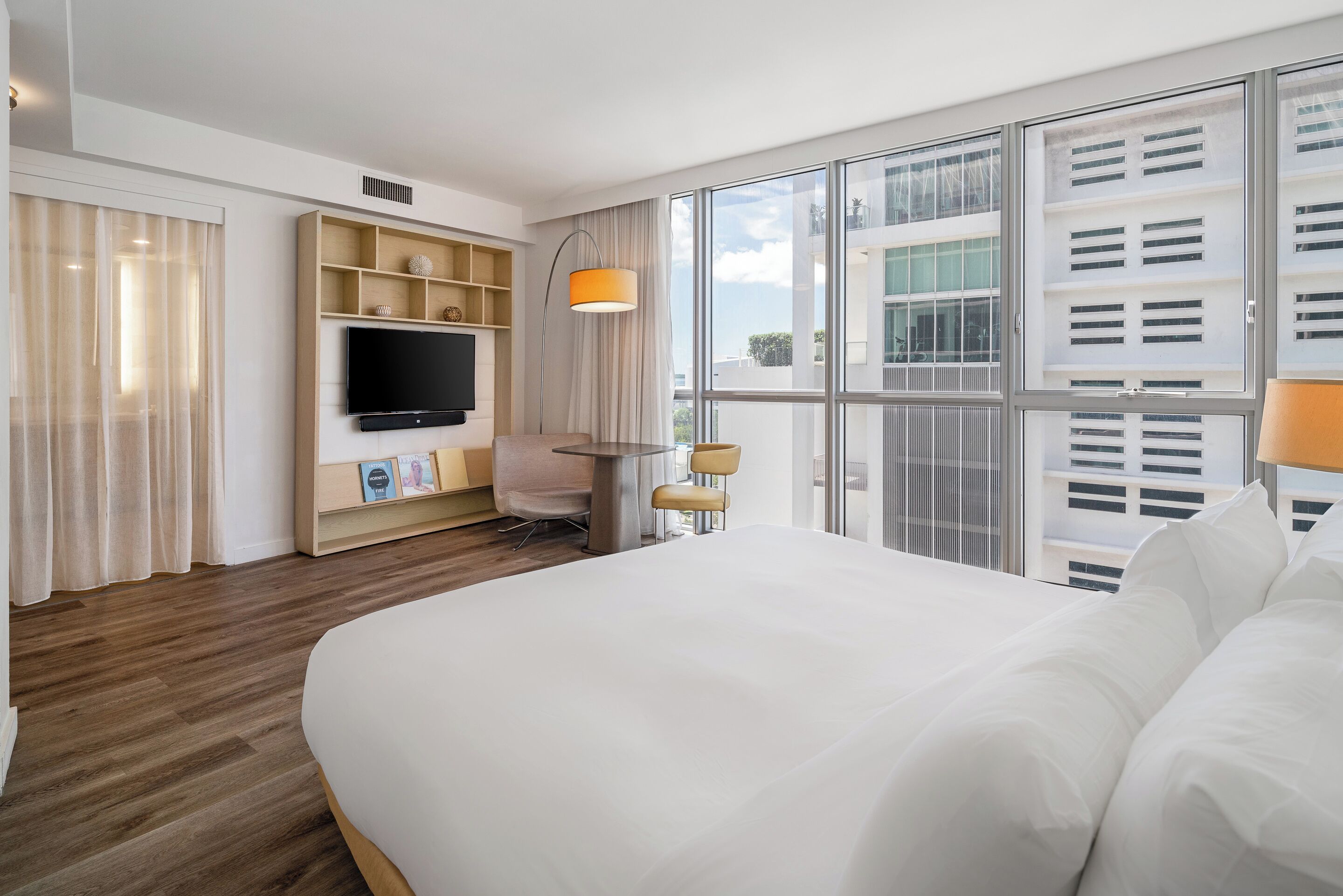 Une chambre d’hôtel avec un grand lit, un téléviseur fixé au mur, un bureau avec une chaise et une fenêtre avec des stores.