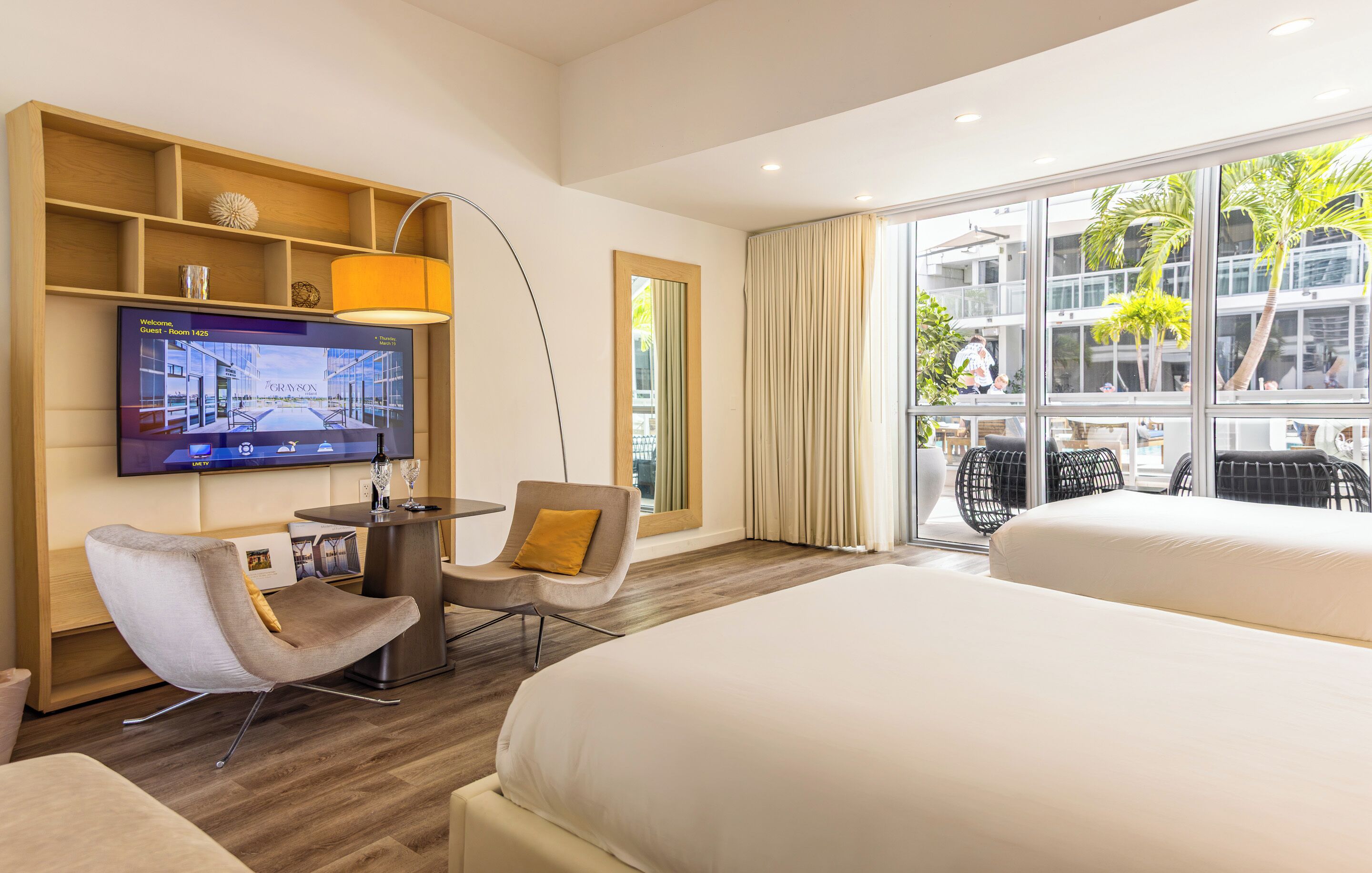Habitación de hotel con televisión, dos camas, escritorio con silla, ventana grande con vista a la ciudad y balcón con mobiliario exterior.