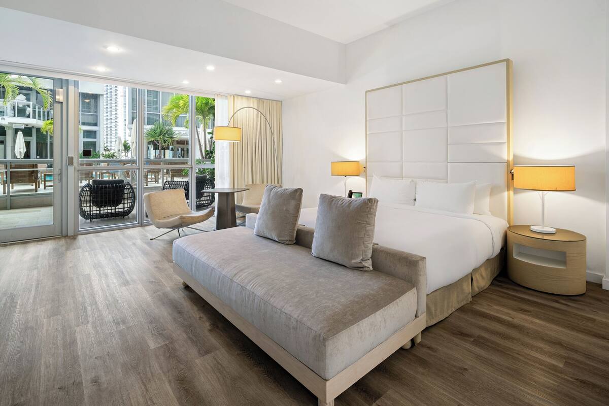 Een moderne hotelkamer met een groot bed, twee nachtkastjes met lampen, een bank en uitzicht op het buitengebied via grote glazen deuren.