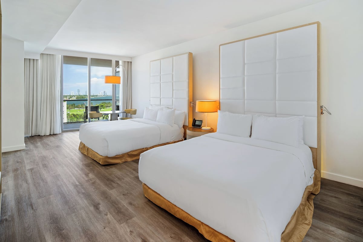 Een hotelkamer met twee bedden, een groot raam met uitzicht op een stadslandschap en groen, en een balkon met een tafel en stoelen.