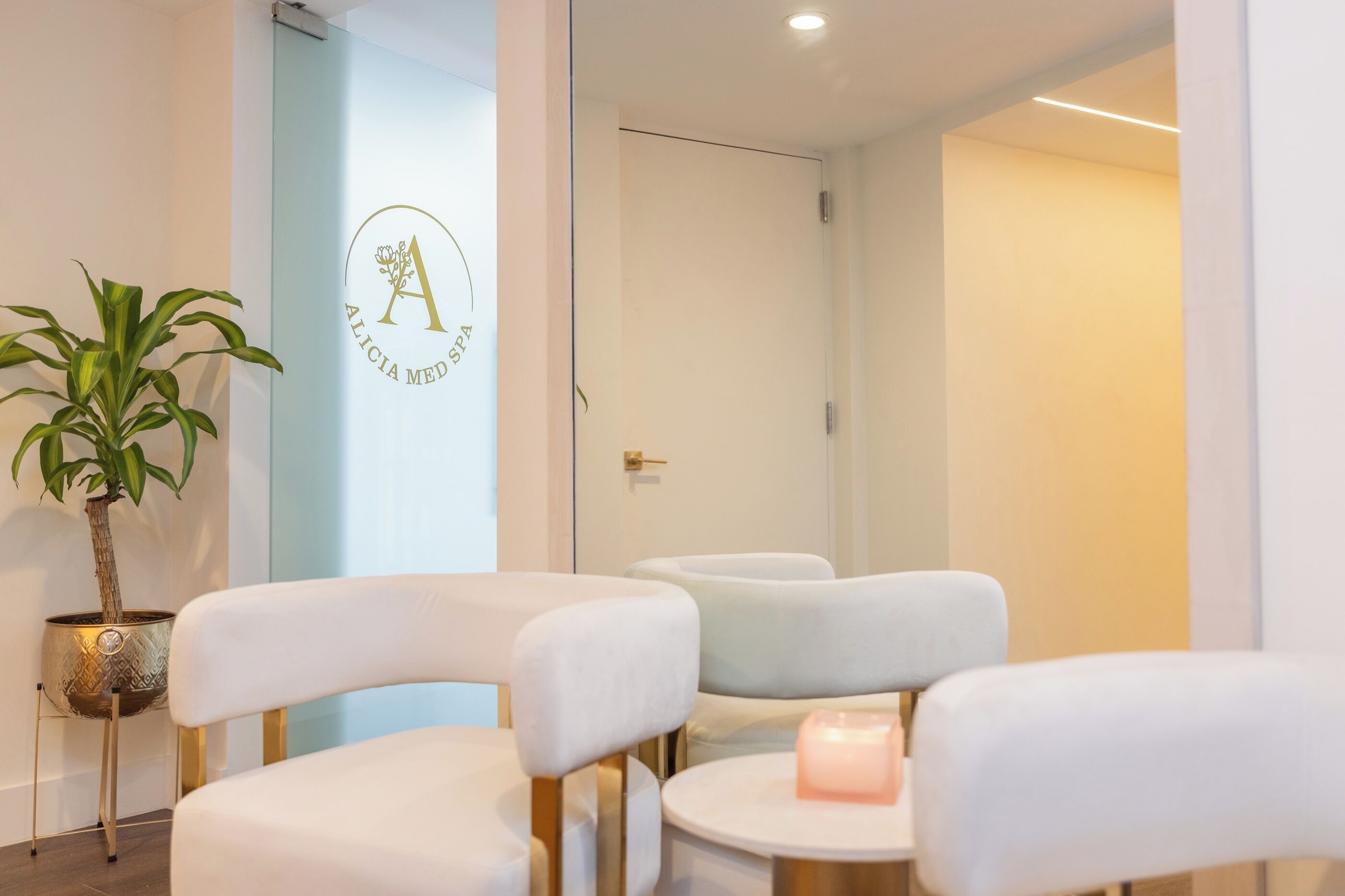 Ein Spa-Empfangsbereich mit Logo an der Wand, einer Topfpflanze und drei weißen Sesseln mit goldfarbenen Beinen.