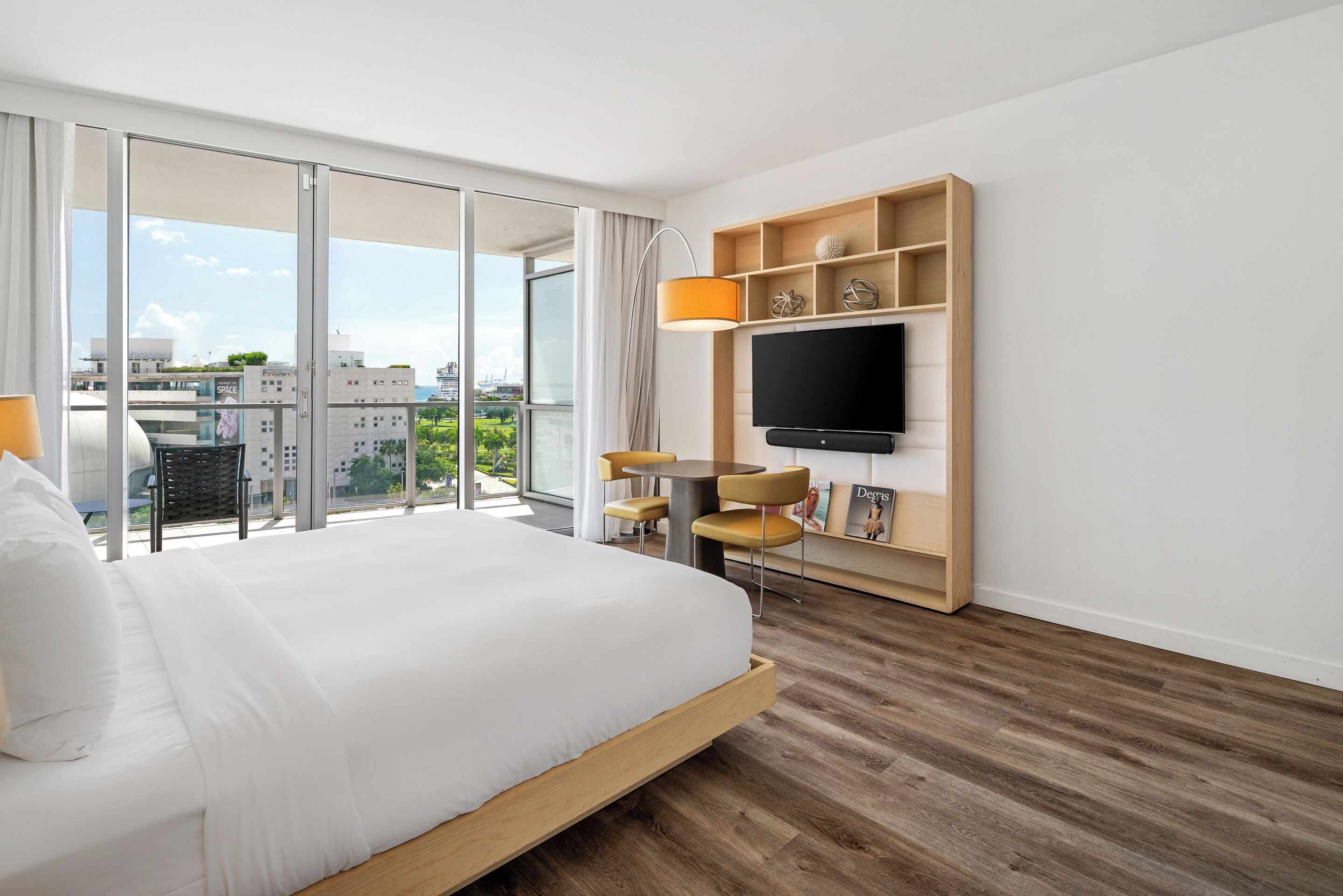 Ett modernt hotellrum med en stor säng, en tv som är monterad på en hylla i trä, en matplats med stolar och utsikt över staden genom stora fönster.