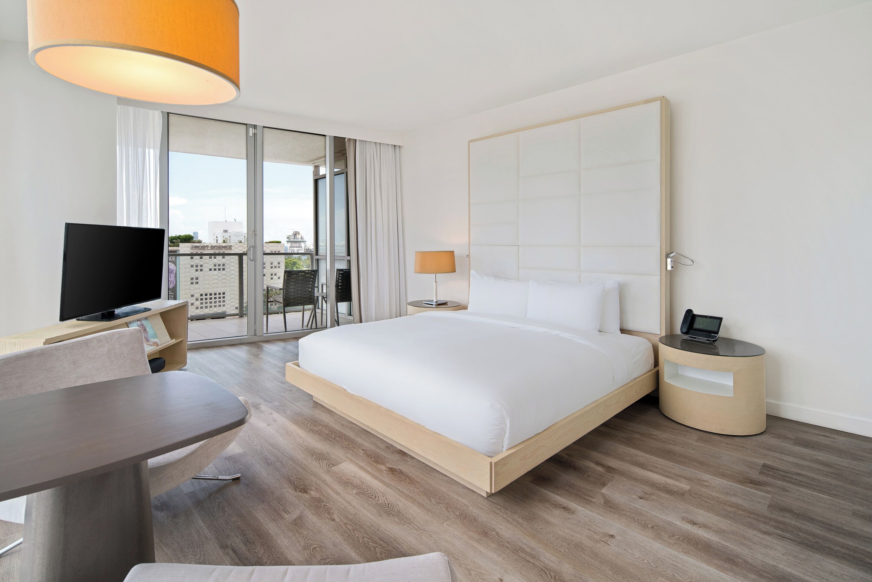 A modern hotel room with a large bed, a desk with a TV, a lamp, and a view of the city through the window.