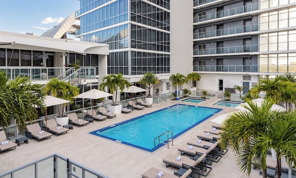 A rooftop pool area with lounge chairs and umbrellas, surrounded by palm trees and modern buildings.