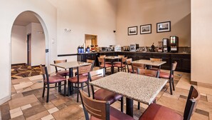 A hotel dining area with multiple tables and chairs, a kitchen area with appliances, and framed pictures on the wall. - Best Western Rambler (Walsenburg)