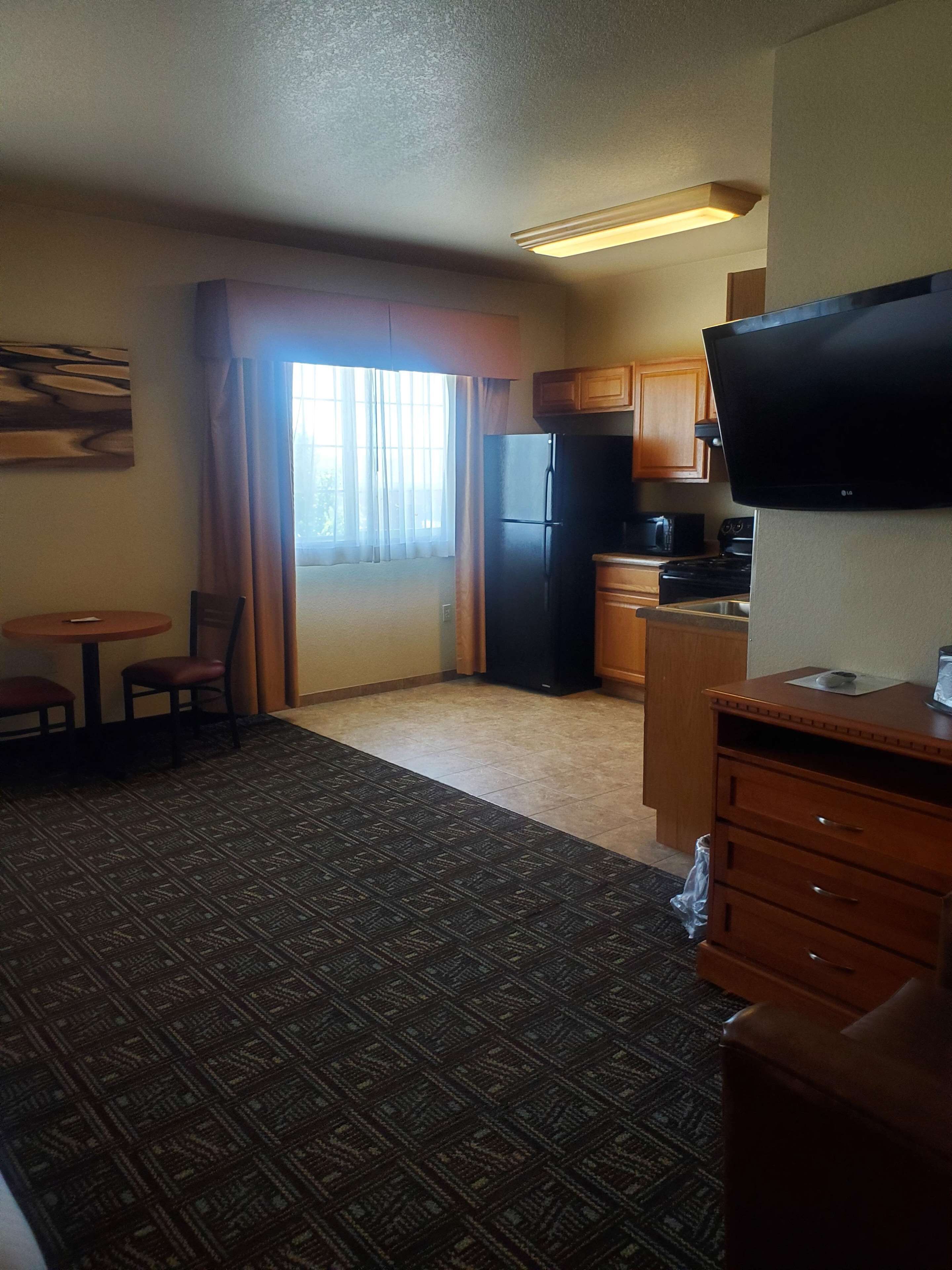 Habitación de hotel compacta con kitchenette, televisor de pantalla plana, zona de comedor con mesa y sillas, y alfombra estampada.