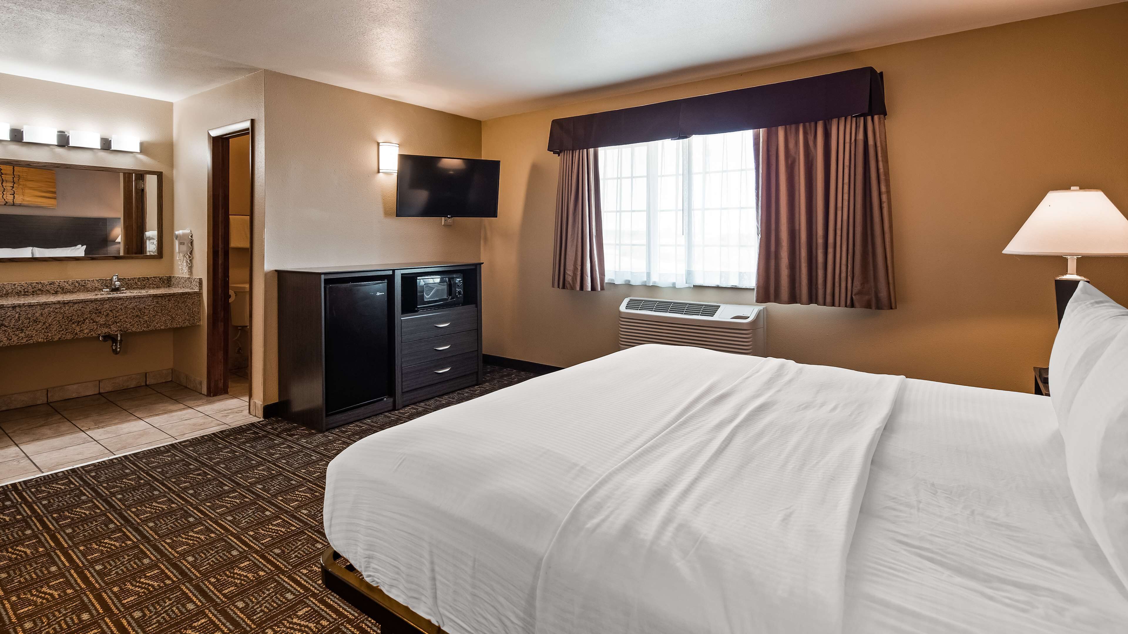 Quarto de hotel com cama, secretária com computador, televisão, janela com cortinas e banheiro com pia e espelho.