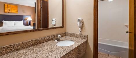 A hotel bathroom with a granite countertop sink, a mirror, a hairdryer, and a bathtub.