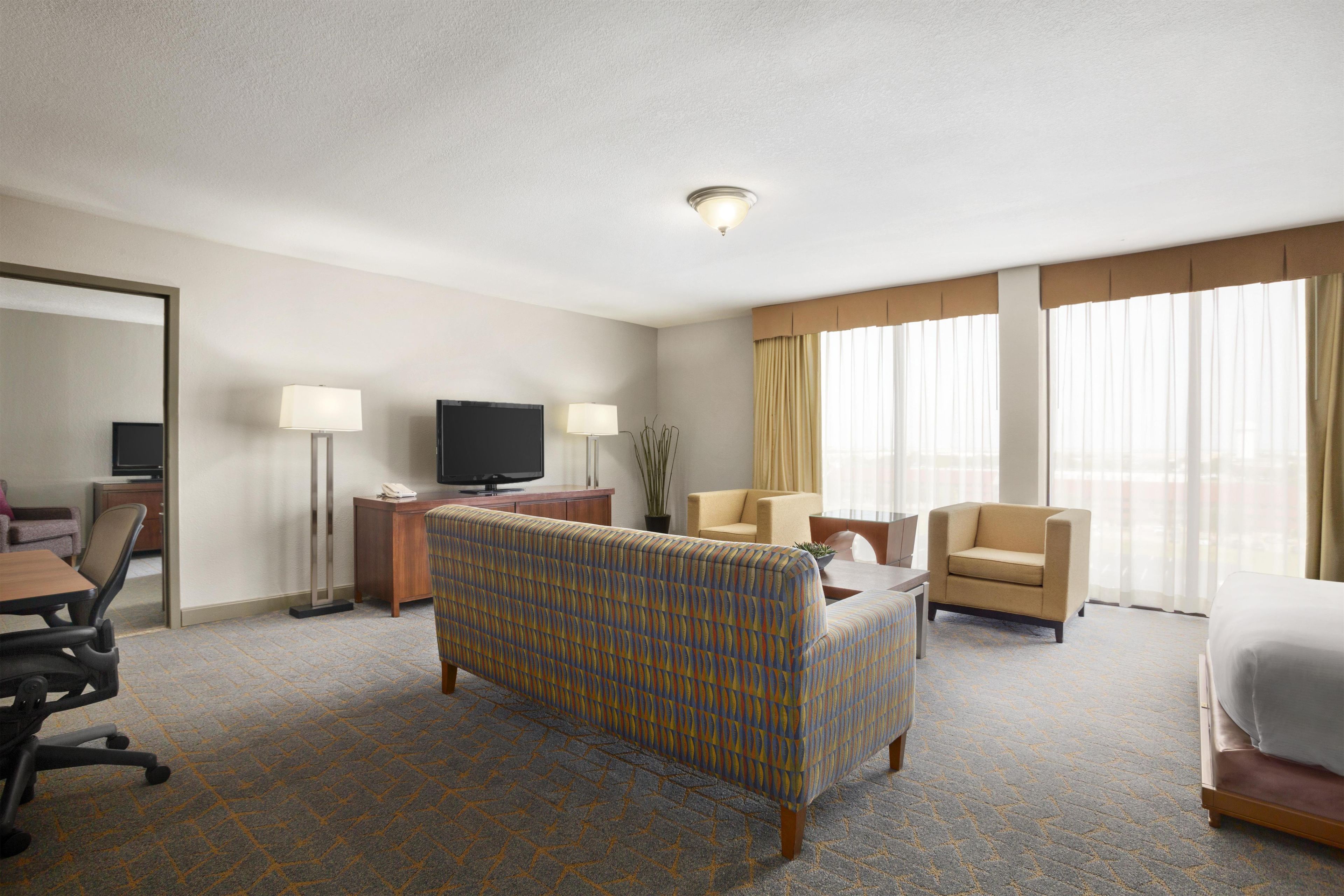 A hotel room with a bed, a sofa, a desk with a TV, a chair, and a large window with curtains.