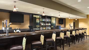 Una zona de bar con una barra alargada, varios taburetes, un televisor montado en la pared y una variedad de botellas y vasos en estantes.