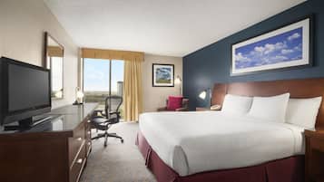 A hotel room with a large bed, a desk with a television, a chair, a desk lamp, a nightstand, and a framed picture on the wall.