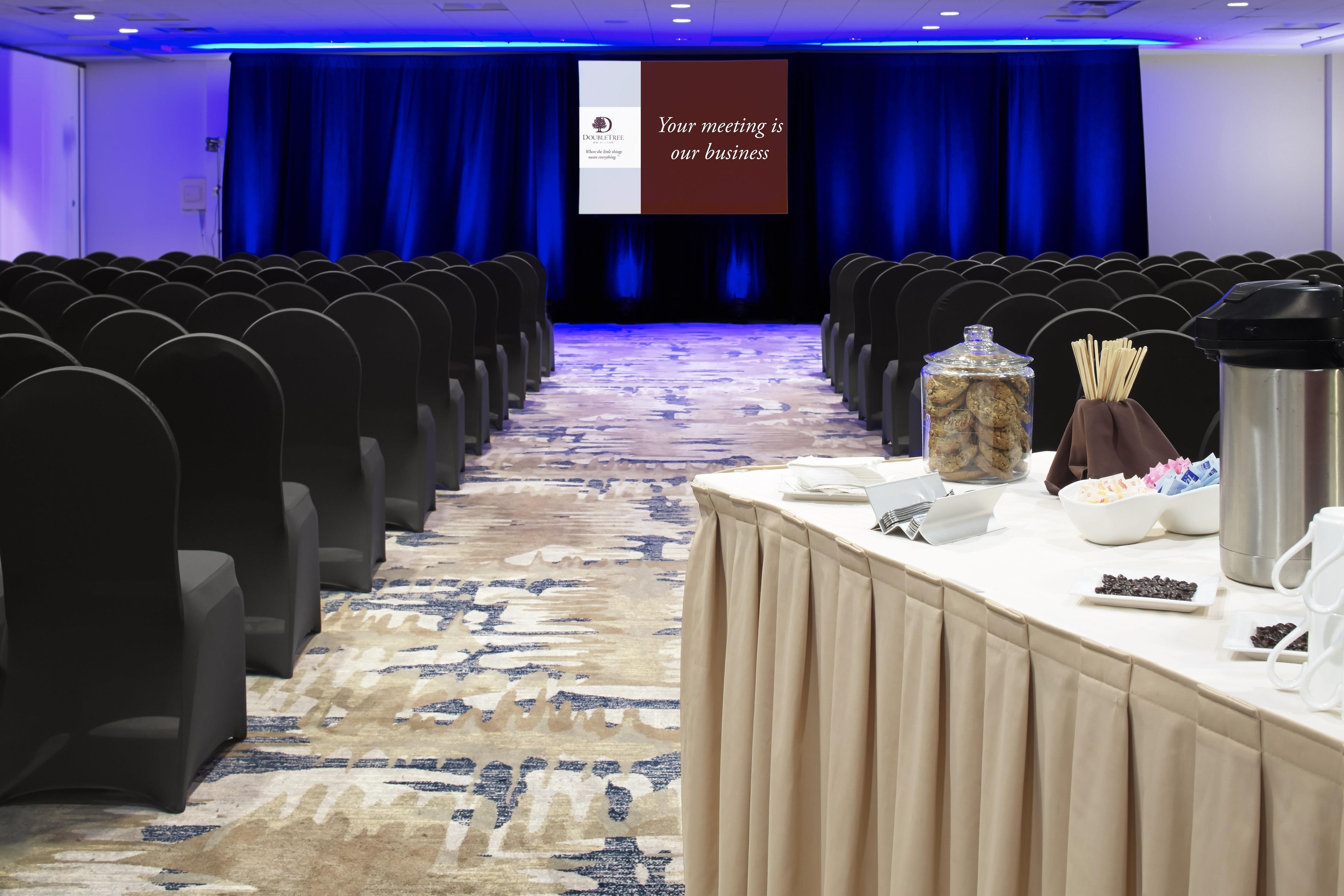 Un salón de conferencias con filas de sillas, una pantalla para presentaciones y una mesa con comida y bebidas.