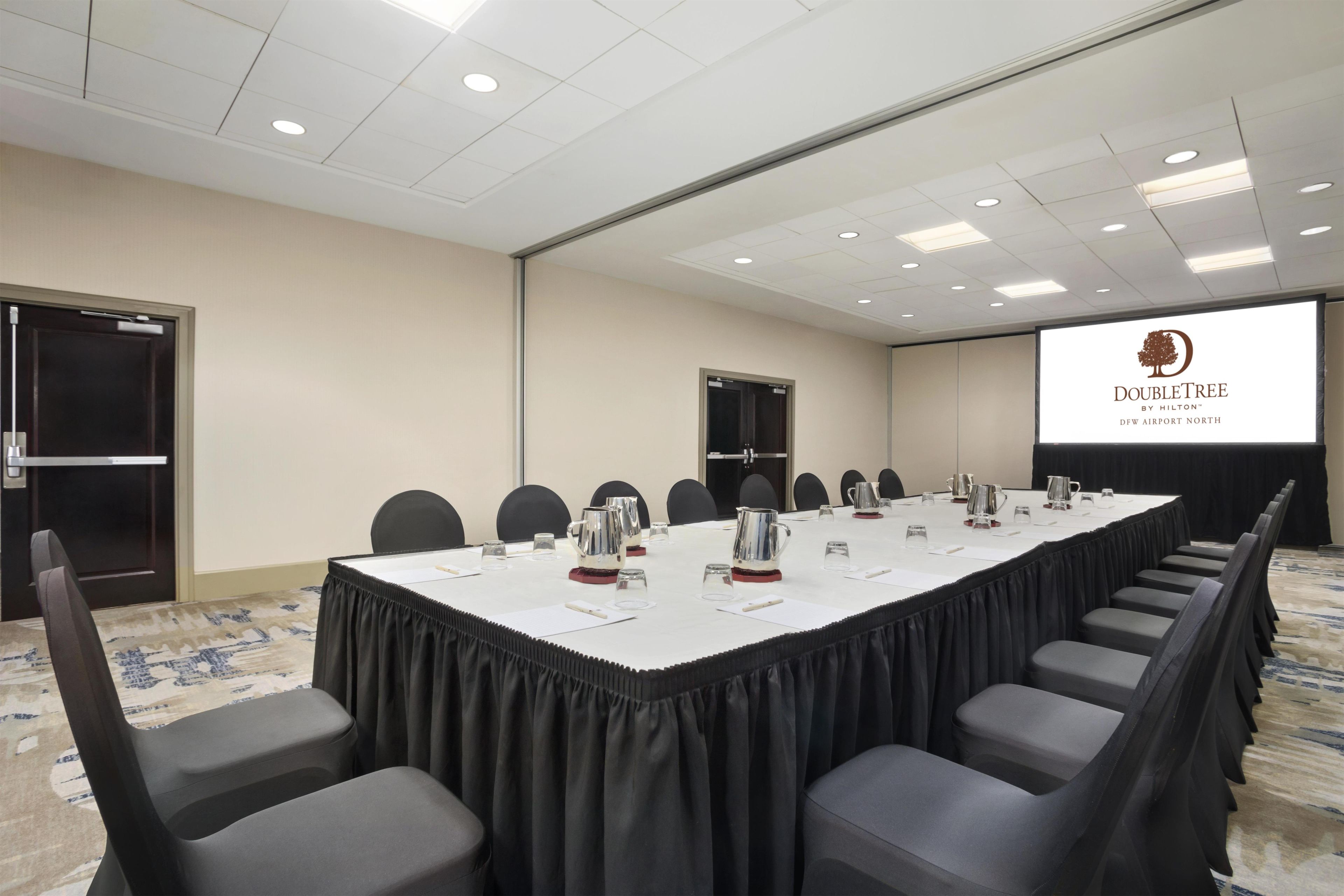 Un salón de conferencias con una larga mesa, sillas, una pantalla de proyector y una puerta.