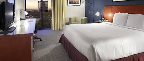 A hotel room with a large bed, a desk, a television, a chair, a nightstand, and a view of the cityscape through the window.