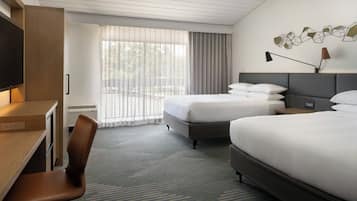 Una habitación de hotel con escritorio, dos camas, televisor y un amplio ventanal con cortinas.