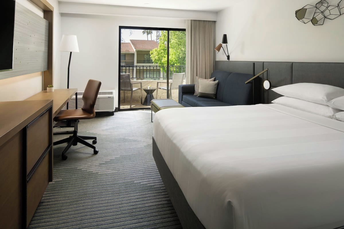 A hotel room with a large bed, a desk, a chair, and a balcony with a view of buildings and trees.