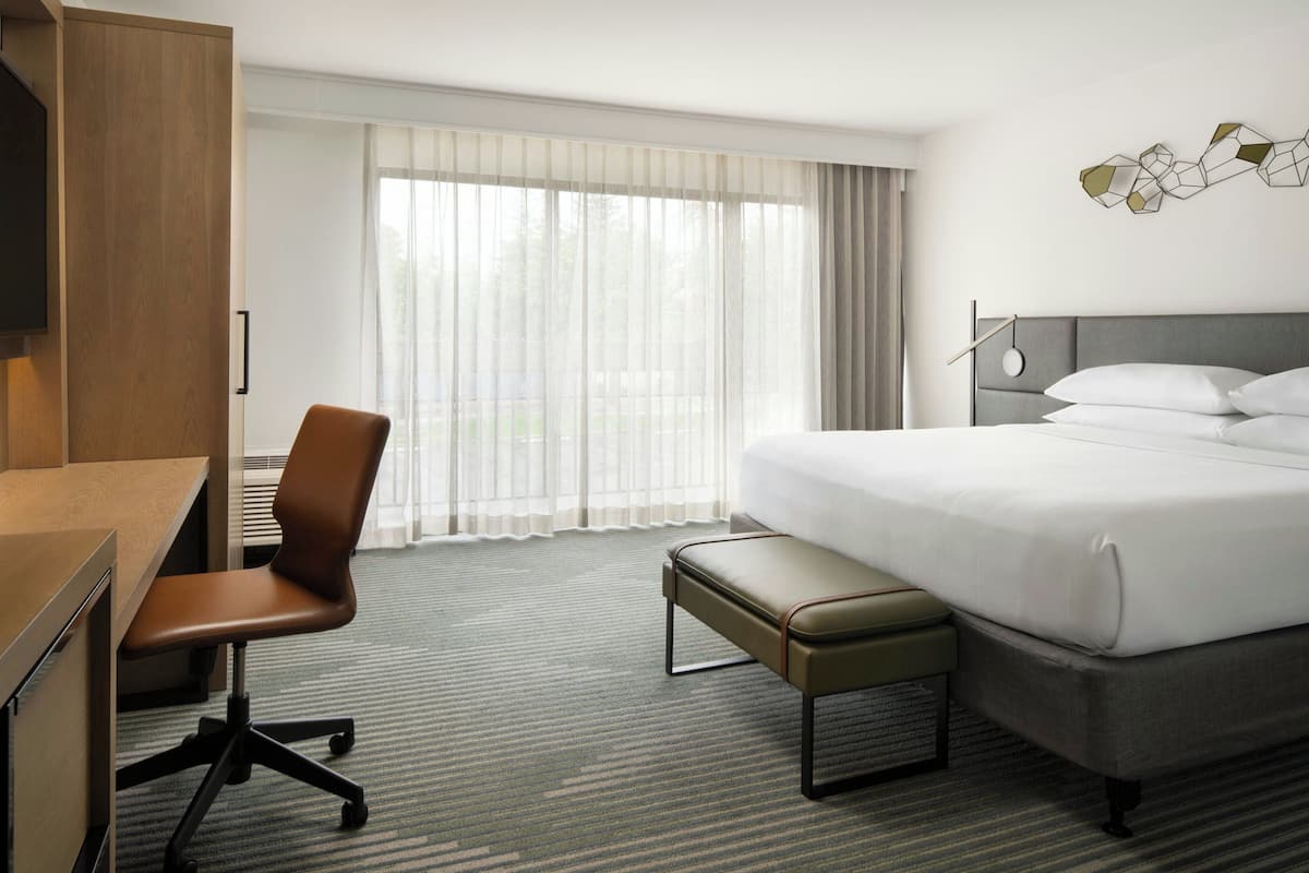 A hotel room with a bed, a desk, a chair, and a large window with sheer curtains.