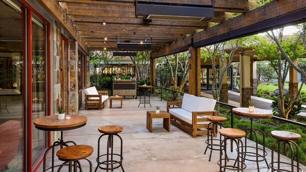 An outdoor seating area with wooden tables and chairs, a sofa, and a bar area with a counter and stools. - Santa Clara Marriott (Santa Clara)