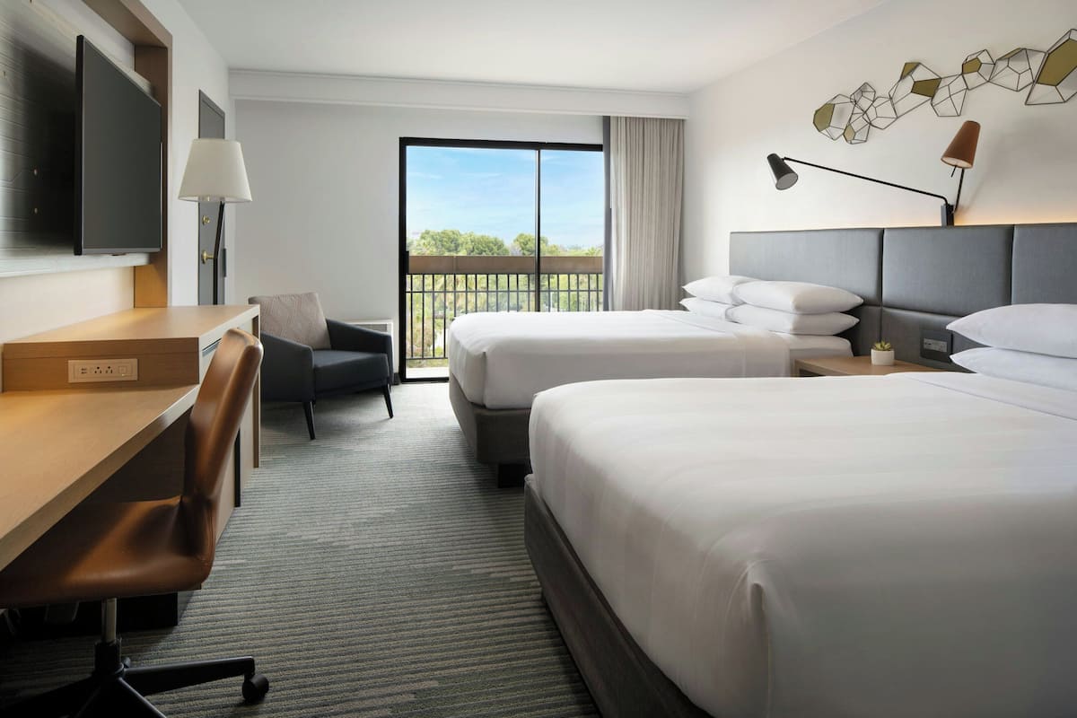 A hotel room with two beds, a desk, a chair, a TV, and a balcony with a view.