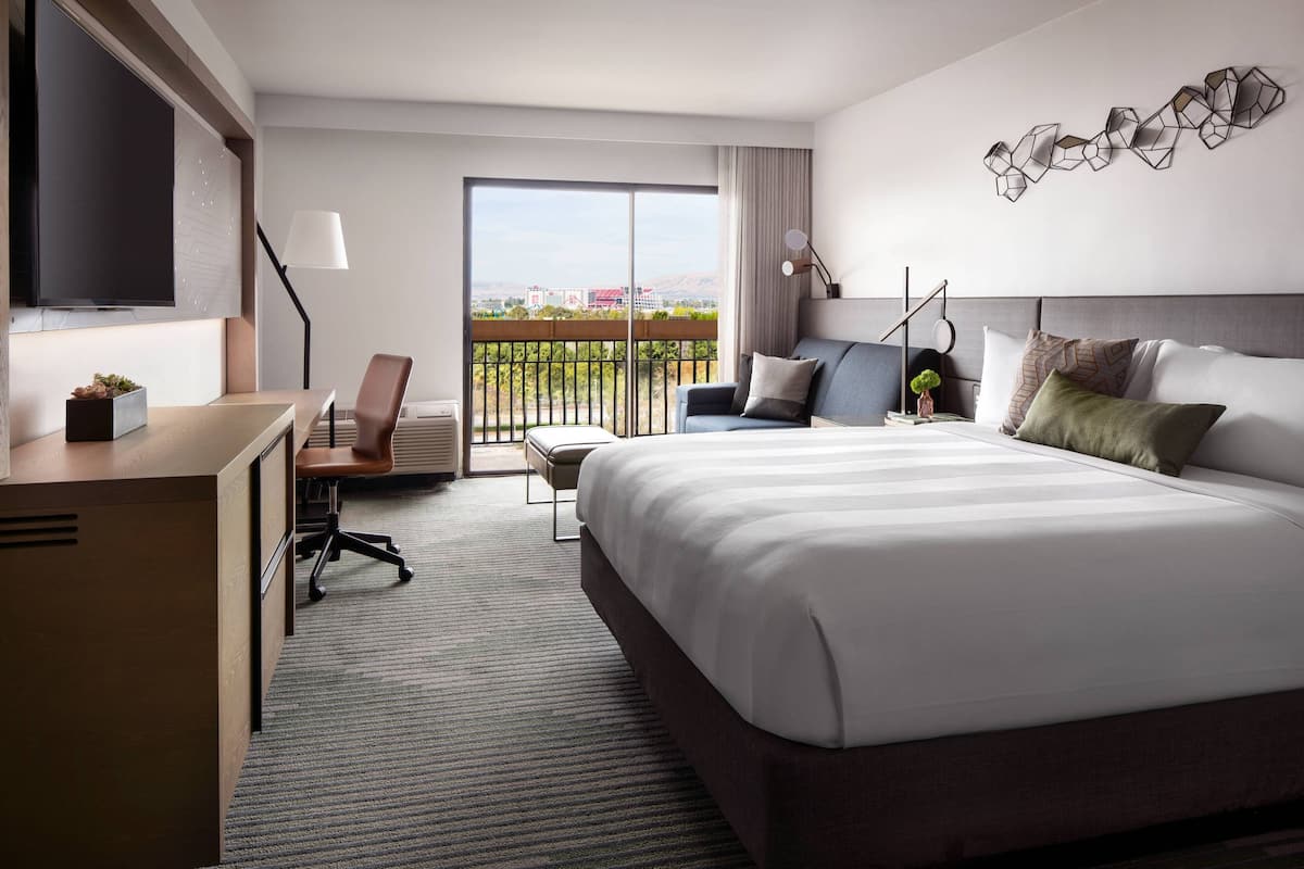 A hotel room with a large bed, a desk with a chair, a TV, and a balcony with a view.