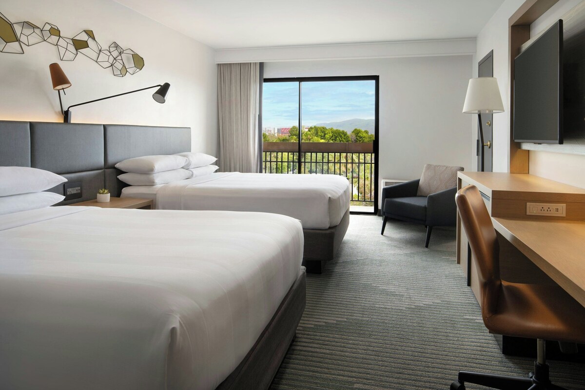 Ein Hotelzimmer mit zwei Betten, einem Schreibtisch, einem Sessel, einem Fernseher und einem Balkon mit Blick auf die Stadt.