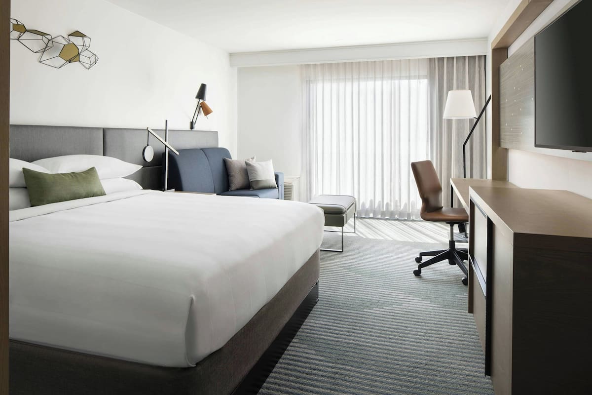 A hotel room with a large bed, a desk, a chair, a TV, and a window with curtains.