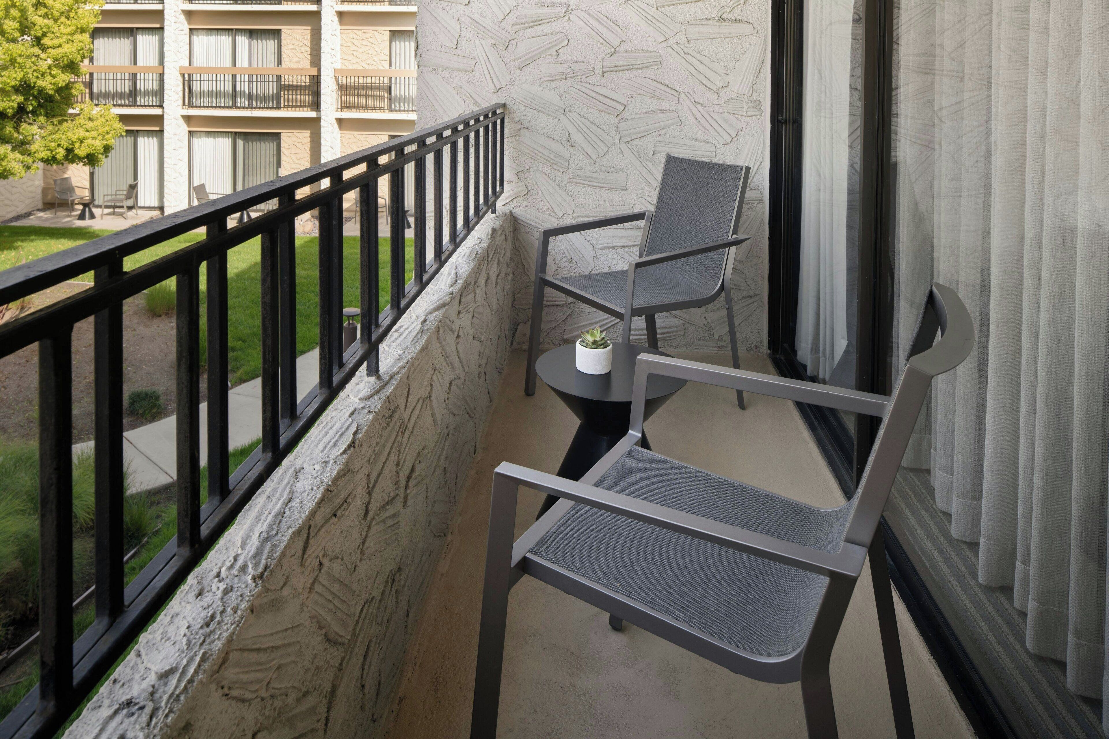 A balcony with two chairs and a small table, a stone wall, and a view of a building and greenery.