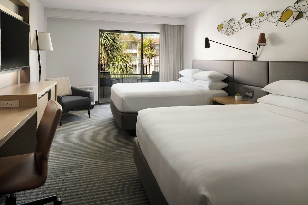 A hotel room with two beds, a desk, a chair, a TV, and a balcony with a view of palm trees.