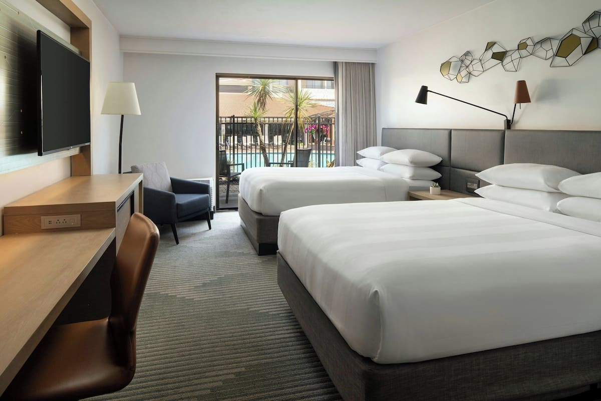 A hotel room with a large bed, a desk, a chair, a TV, and a balcony with a pool view.