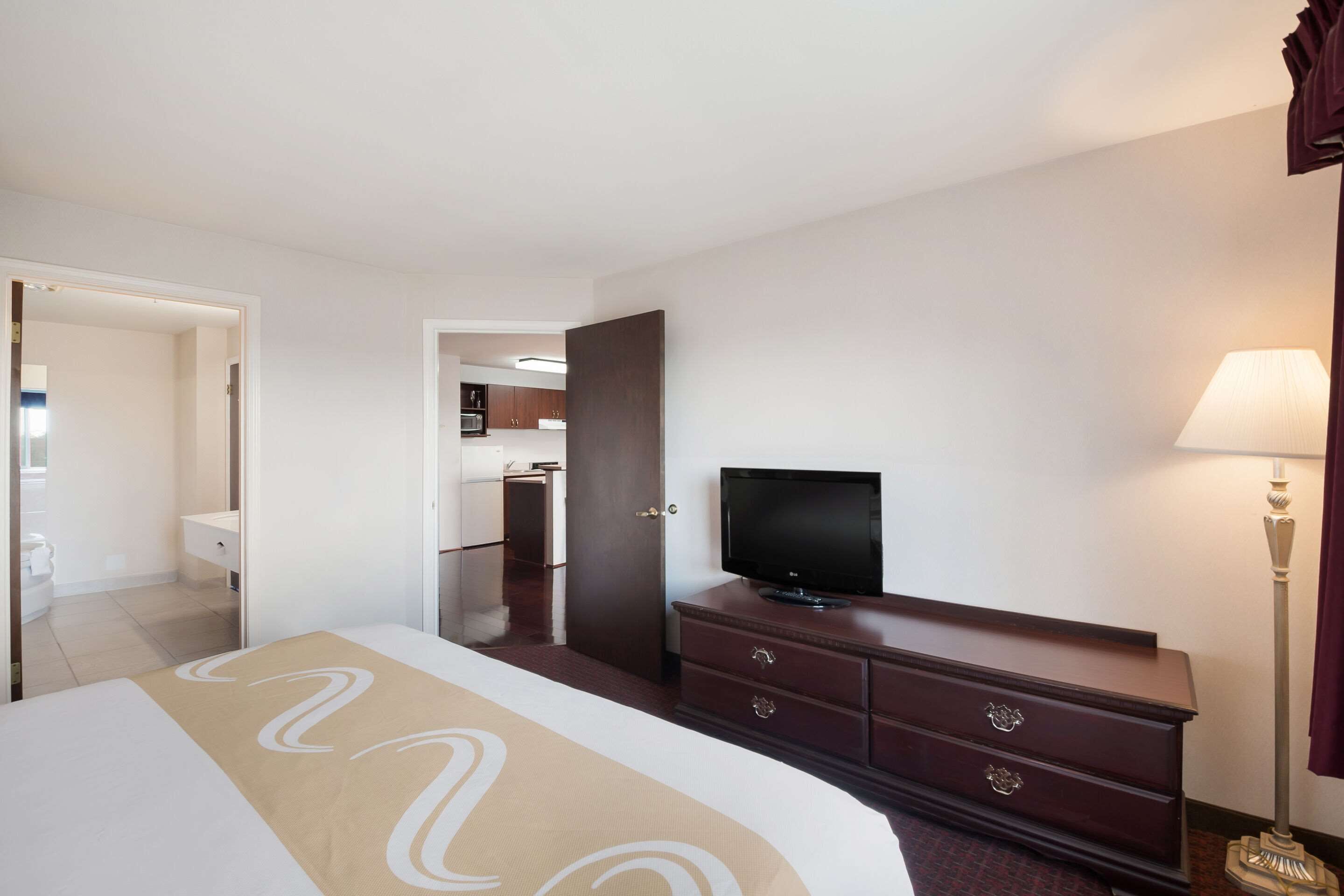 Hotelkamer met een bed, een televisie, een dressoir en een lamp. Er is een deur naar een badkamer en een kitchenette in de achtergrond.