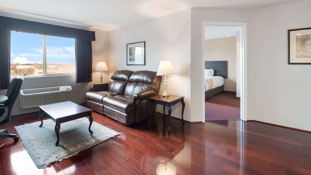 A hotel room with a leather sofa, a small table, a desk, and a bed. There is a window with curtains, a picture on the wall, and a radiator. - Quality Inn Grand Suites Bellingham (Bellingham)