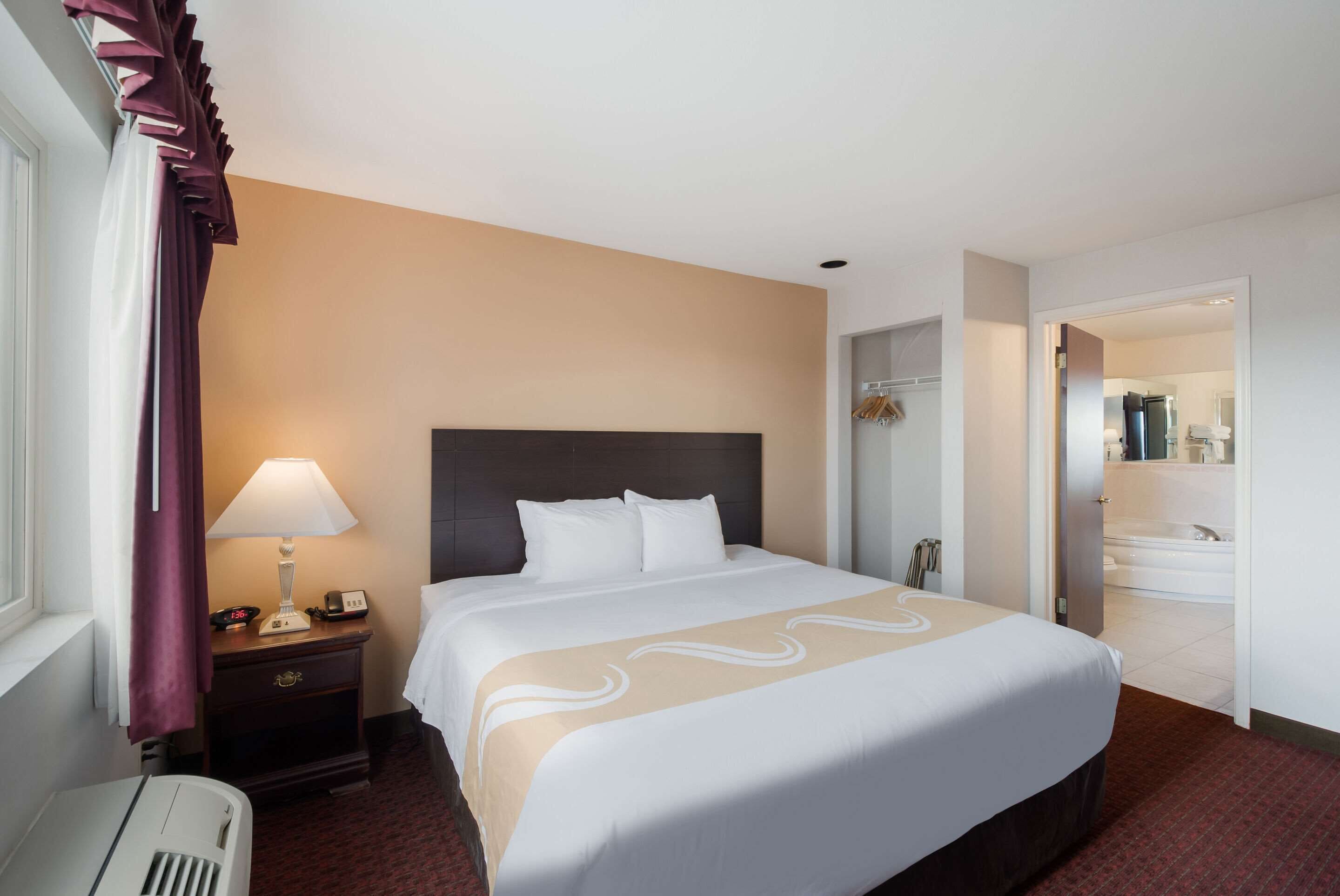 Ein Hotelzimmer mit einem großen Bett, einem Nachttisch mit Lampe, einem kleinen Tischchen und Blick ins Badezimmer.