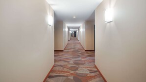 A hotel hallway with patterned carpet, light-colored walls, and wall-mounted light fixtures. - Quality Inn Grand Suites Bellingham (Bellingham)