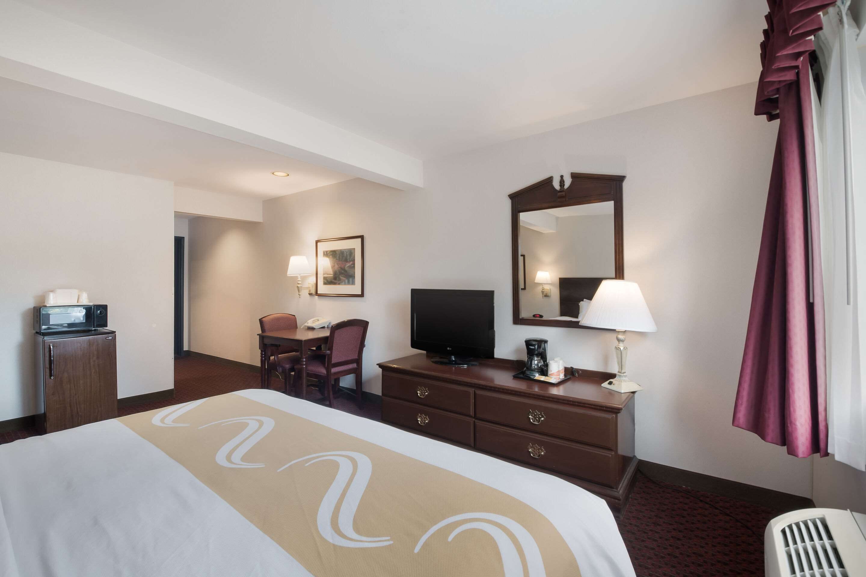 A hotel room with a bed, a desk, a television, a mirror, a lamp, and a window with curtains.