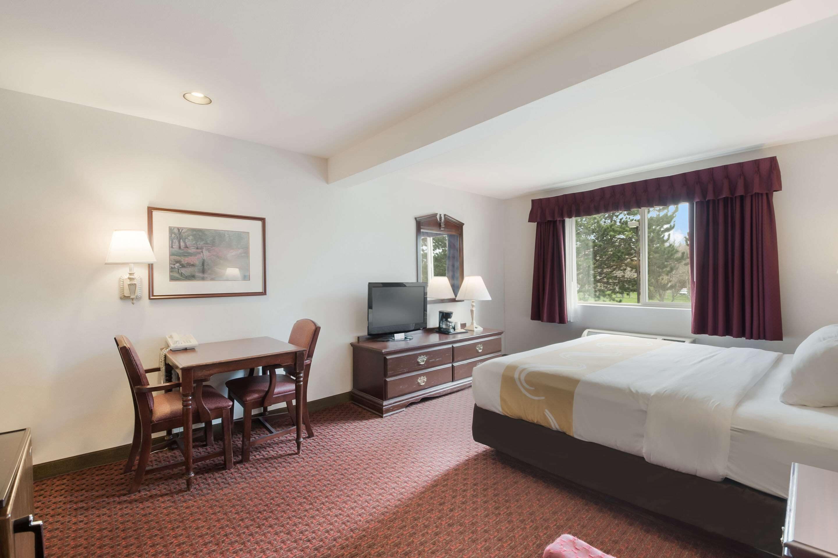 A hotel room with a bed, a desk with a chair, a television, a dresser, and a window with curtains.