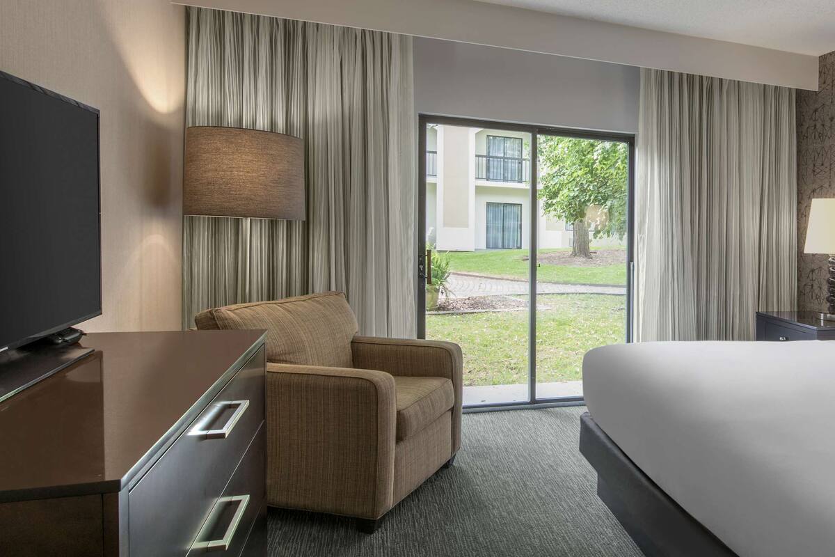 A hotel room with a bed, a desk, a chair, a TV, and a view of the outside.