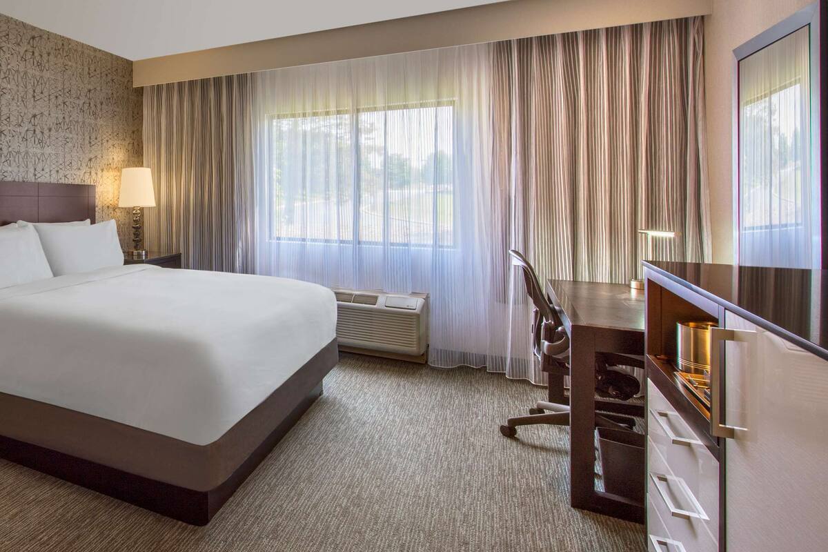 A hotel room with a large bed, two bedside lamps, a wardrobe, a desk with a chair, and a large window with curtains.