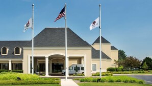A building with a beige facade, a dark roof, and a central entrance with columns. There are two flags on flagpoles in front. - DoubleTree by Hilton Hotel Chicago Wood Dale - Elk Grove (Wood Dale)