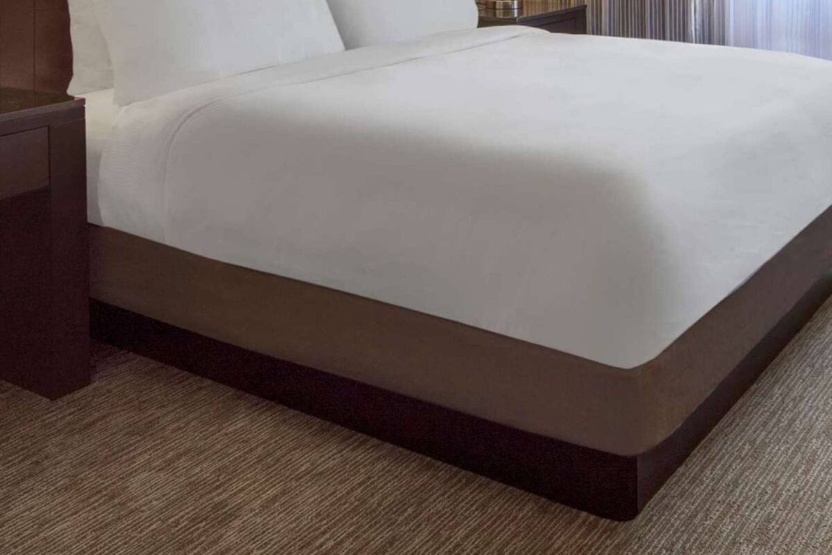 A large bed with white linens, a dark headboard, and a patterned wallpaper. There is a nightstand with a lamp and a window with curtains.