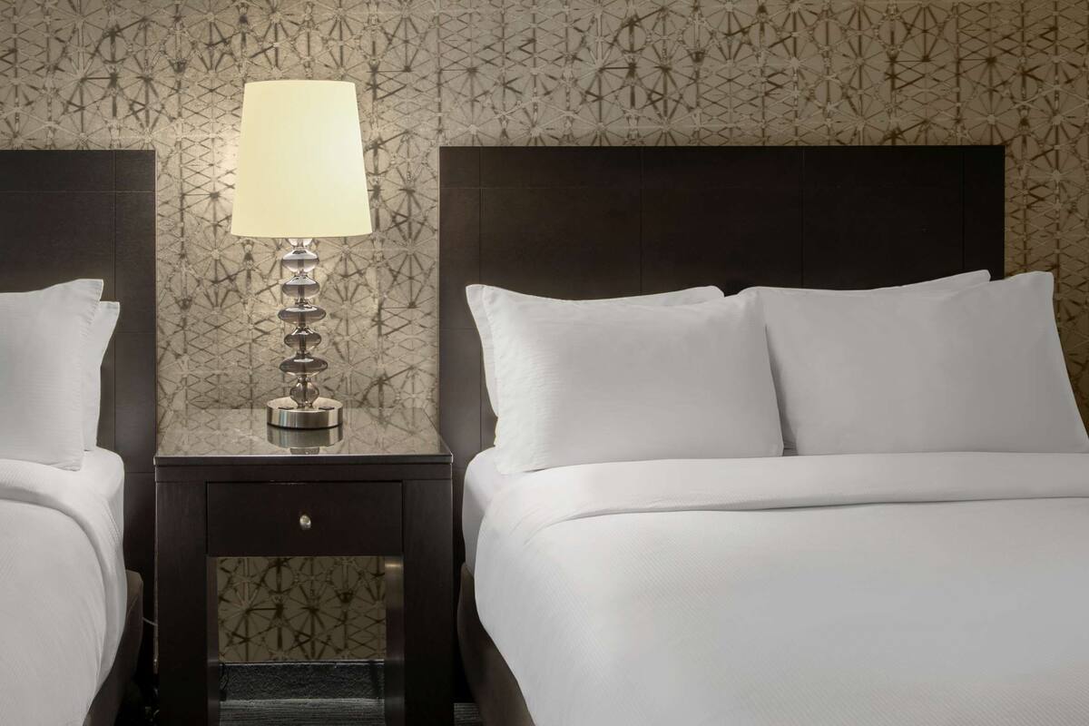 A hotel room with two beds, a nightstand with a lamp, and patterned wallpaper.