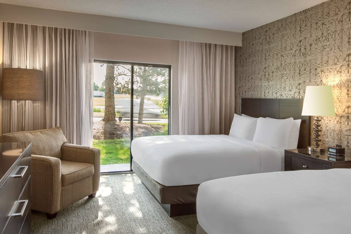 A hotel room with two beds, a desk with a TV, a chair, and a view of the outdoors.