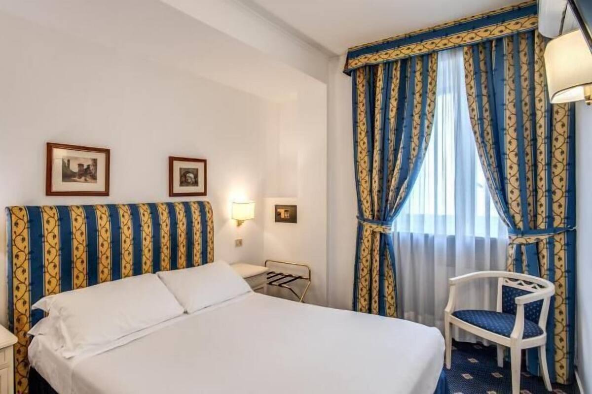 Una habitación de hotel con cama, lámparas de noche, una silla y cortinas.
