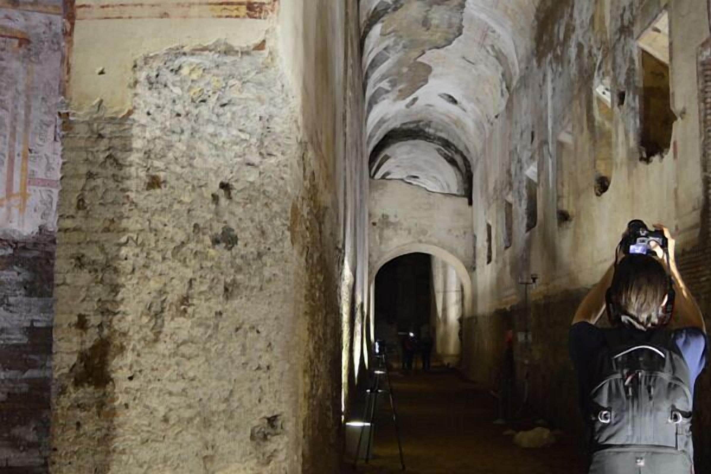 A person taking a photo in an ancient, dimly lit corridor with stone walls and an arched ceiling.