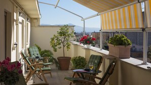 Un balcón con sillas, una mesa, plantas en macetas y vistas a la ciudad y las montañas.