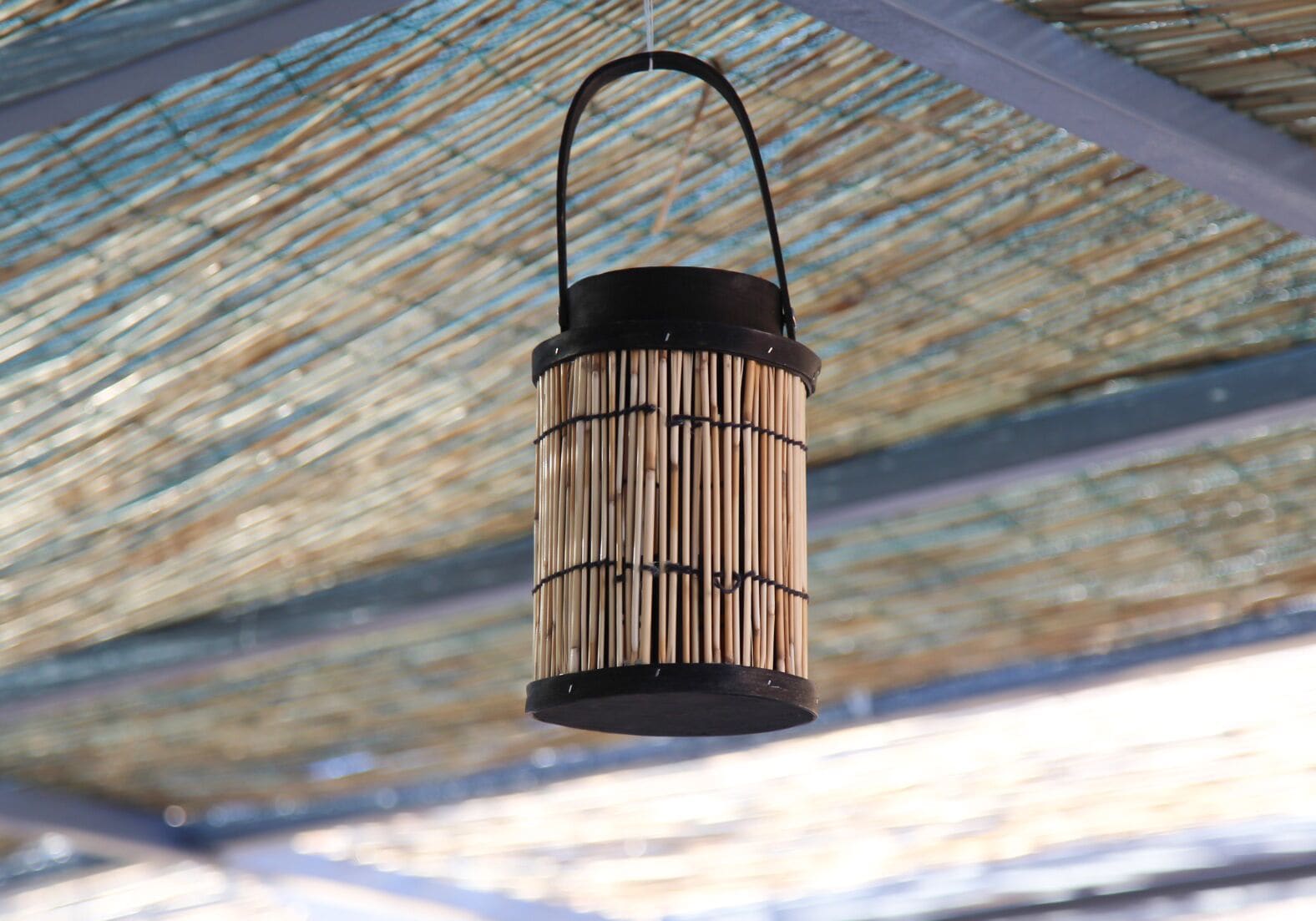 A hanging bamboo lantern with a black handle, suspended from a ceiling with a woven pattern.