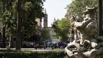 A park with a stone sculpture, trees, and a distant building with a tower.