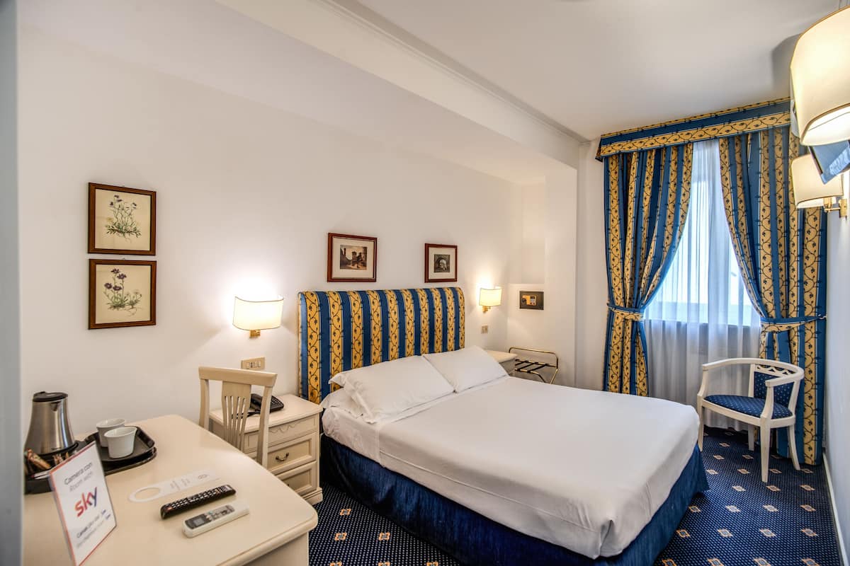 Una habitación de hotel con cama, dos mesitas de noche, un escritorio con silla, lámpara y ventana con cortinas.